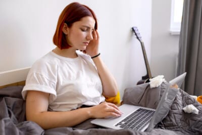 Woman sitting on a bed using a laptop while holding her temple