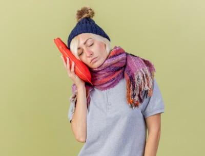 Woman wearing a scarf and a knitted hat holding a red hot water bottle to her face