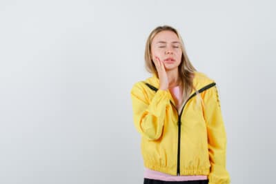 Woman in a yellow jacket holding her cheek with a pained expression