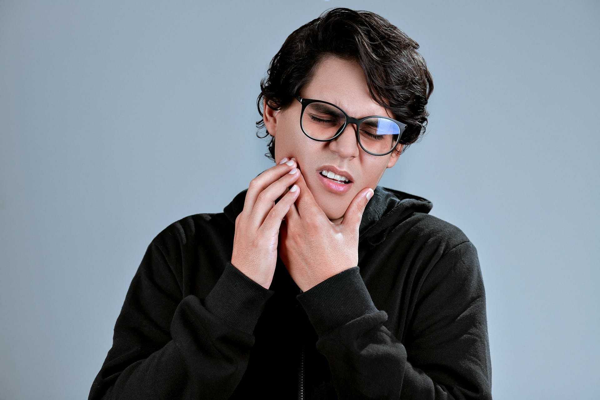Young person wearing glasses and holding both hands to their jaw with a pained expression