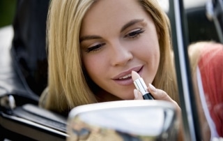 Young woman putting on lip stick in a car mirror